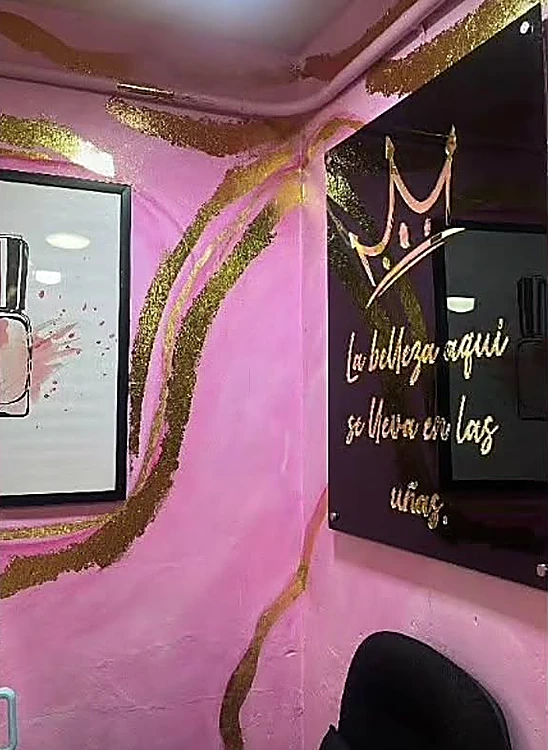 Letrero negro con corona dorada que dice: La belleza aquí se lleva en las uñas, sobre el muro rosa del salón